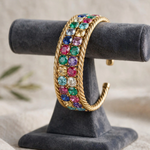 Illustrative styling image of a gold-tone twist cuff bracelet with multi-colour stones on a display stand