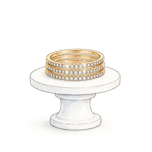 Illustrative dainty gold-toned stackable bands with clear stones arranged on a white pedestal